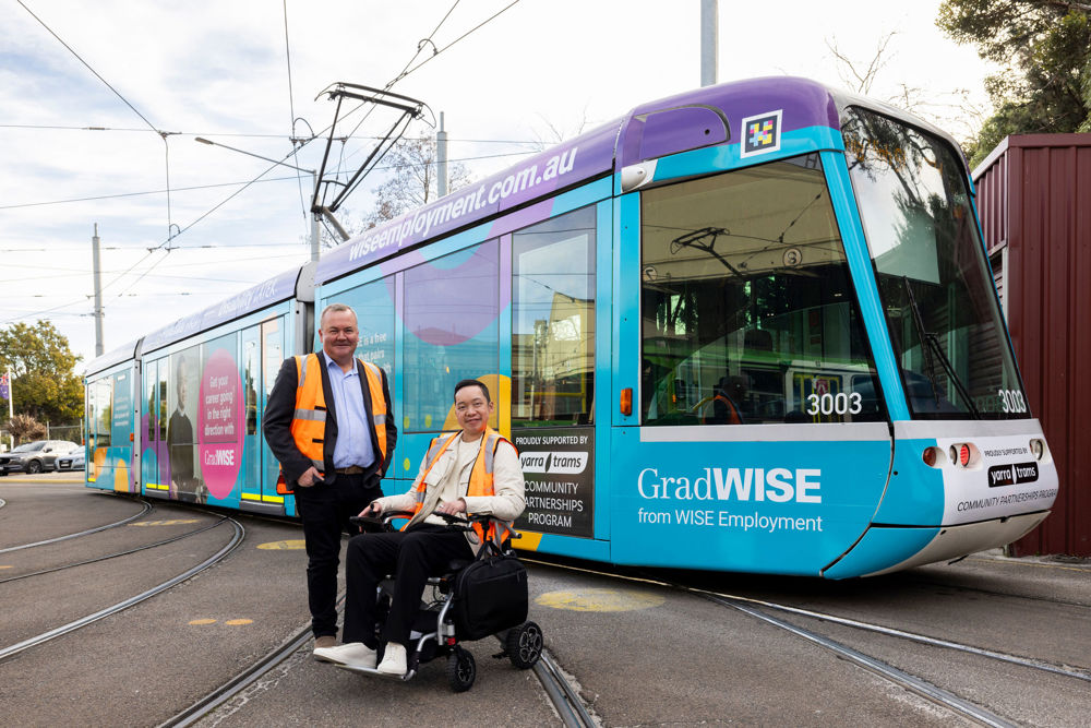Two men in high vis vests pose infront of a C1 tram, one man is standing next to the other who is in a motorised wheelchair. The words